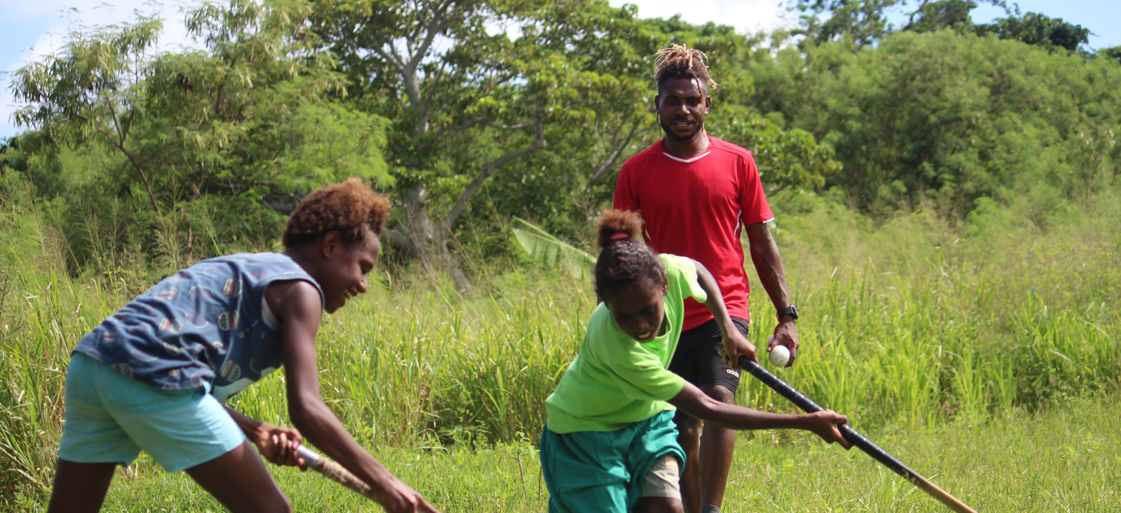 Two girls play hockey on a grass field as a male coach watches on in the background