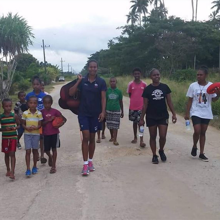 A woman carrying a bag of AFL balls on her shoulder walking down a road with children and a man walking beside her 