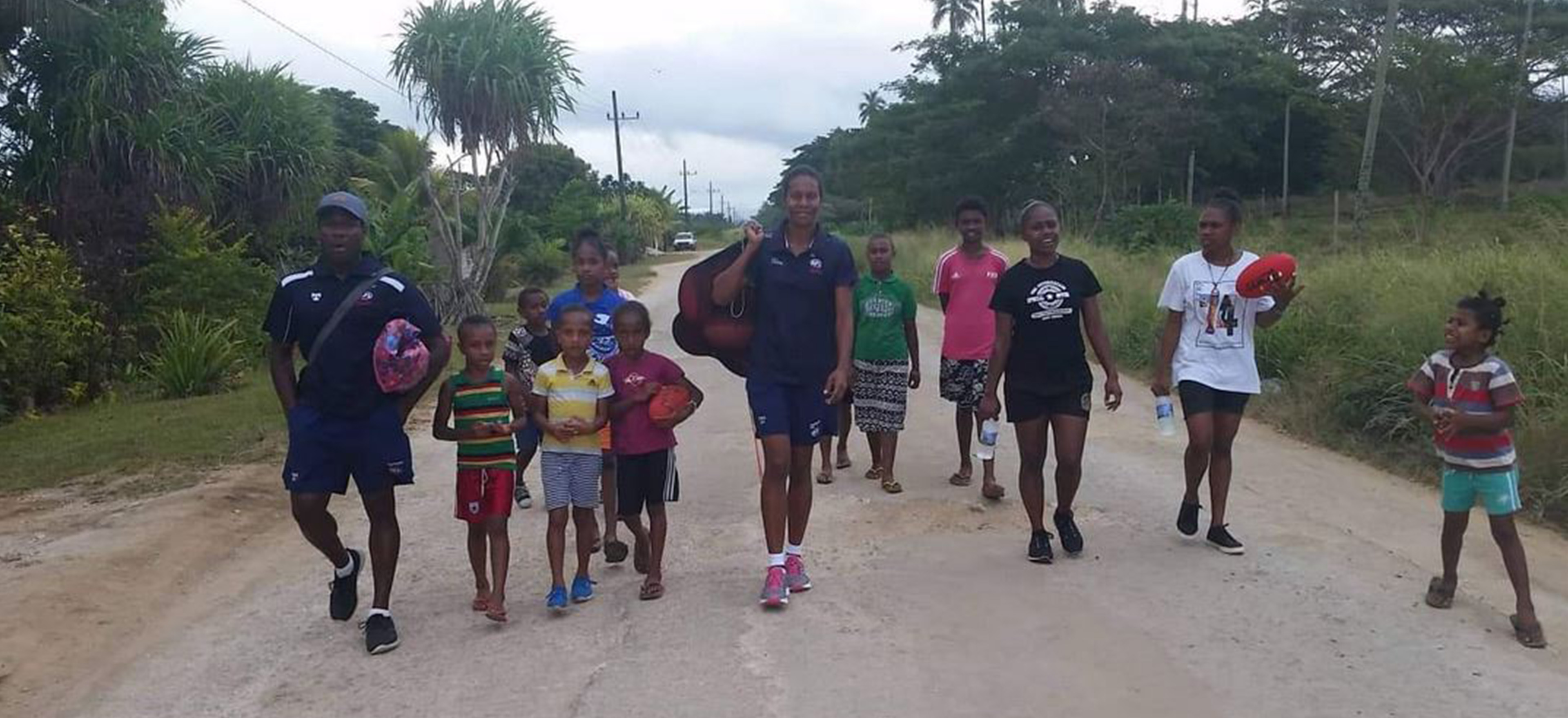 A woman carrying a bag of AFL balls on her shoulder walking down a road with children and a man walking beside her