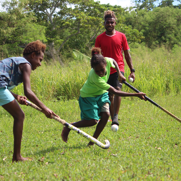 Two girls play hockey on a grass field as a male coach watches on in the background 